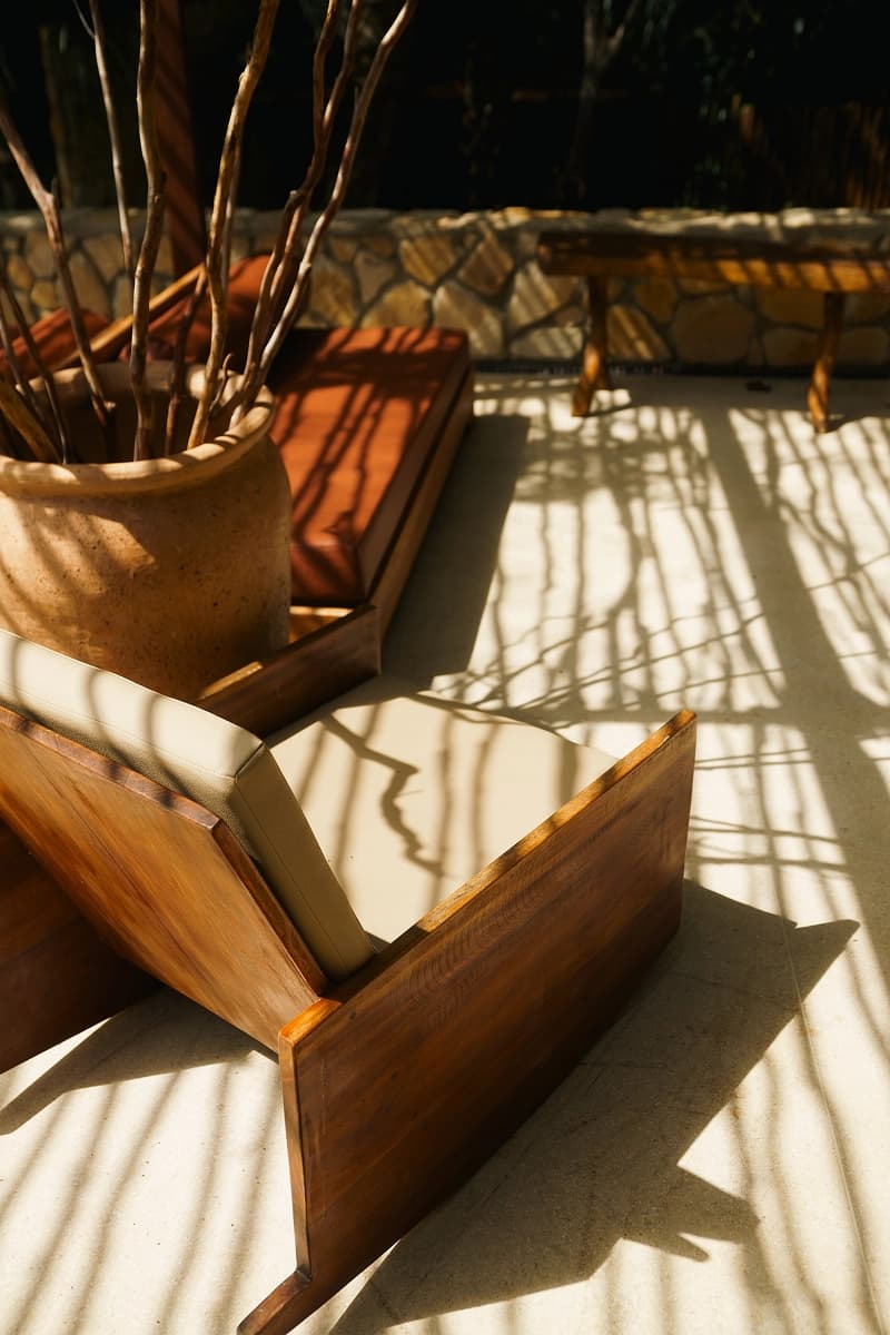 Área externa com poltronas de madeira, almofadas claras e sombras desenhadas por galhos